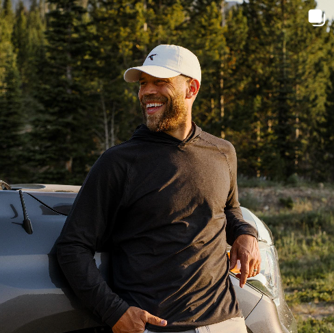 Man leaning against a truck wearing GenTeal Black Heathered Long Sleeve Hooded T-Shirt and White Logo Hat